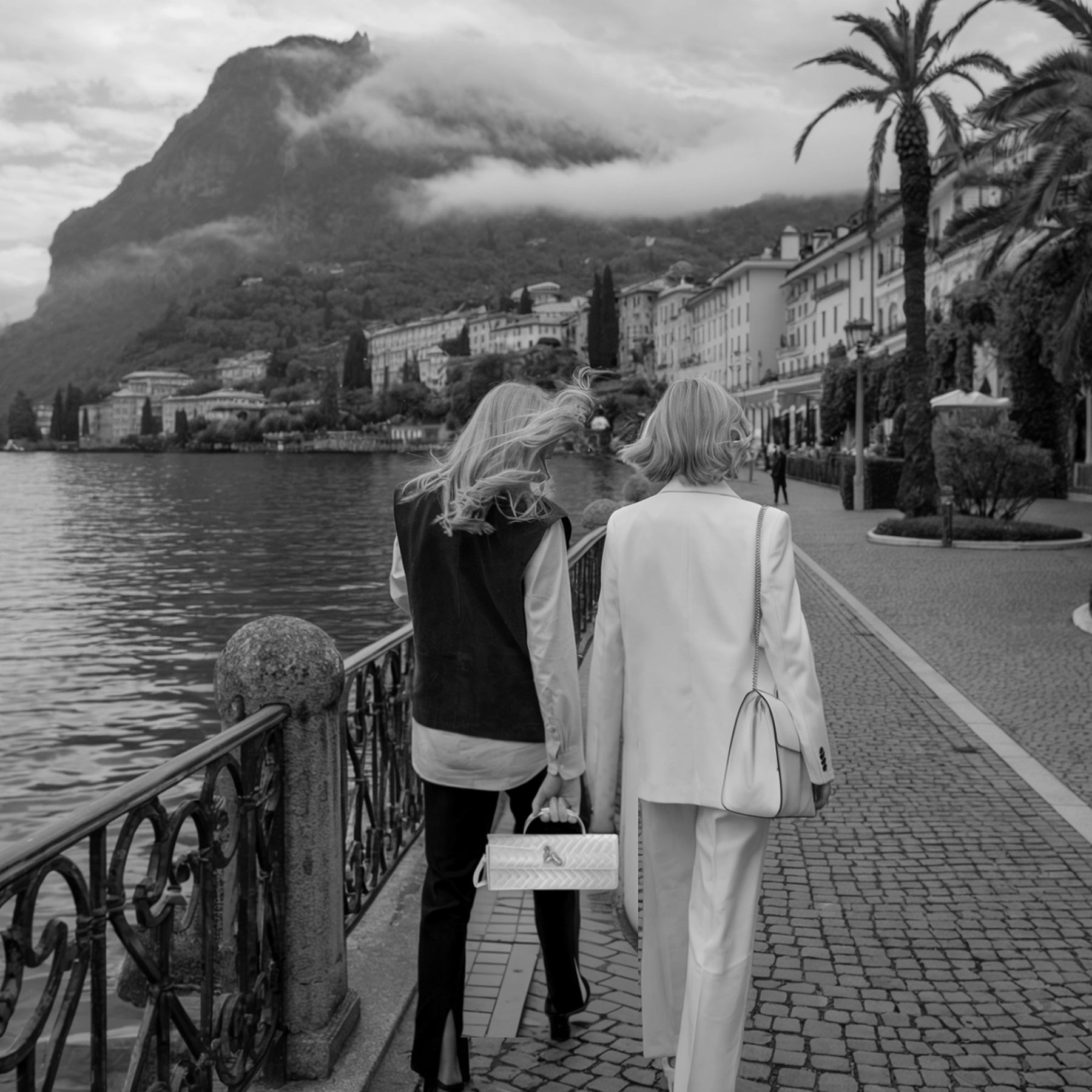 Two women walking along a lakeside promenade with mountains and buildings in the background.
