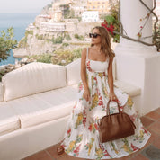 Woman in a floral dress holding a brown handbag, sitting on a white couch with a scenic background.