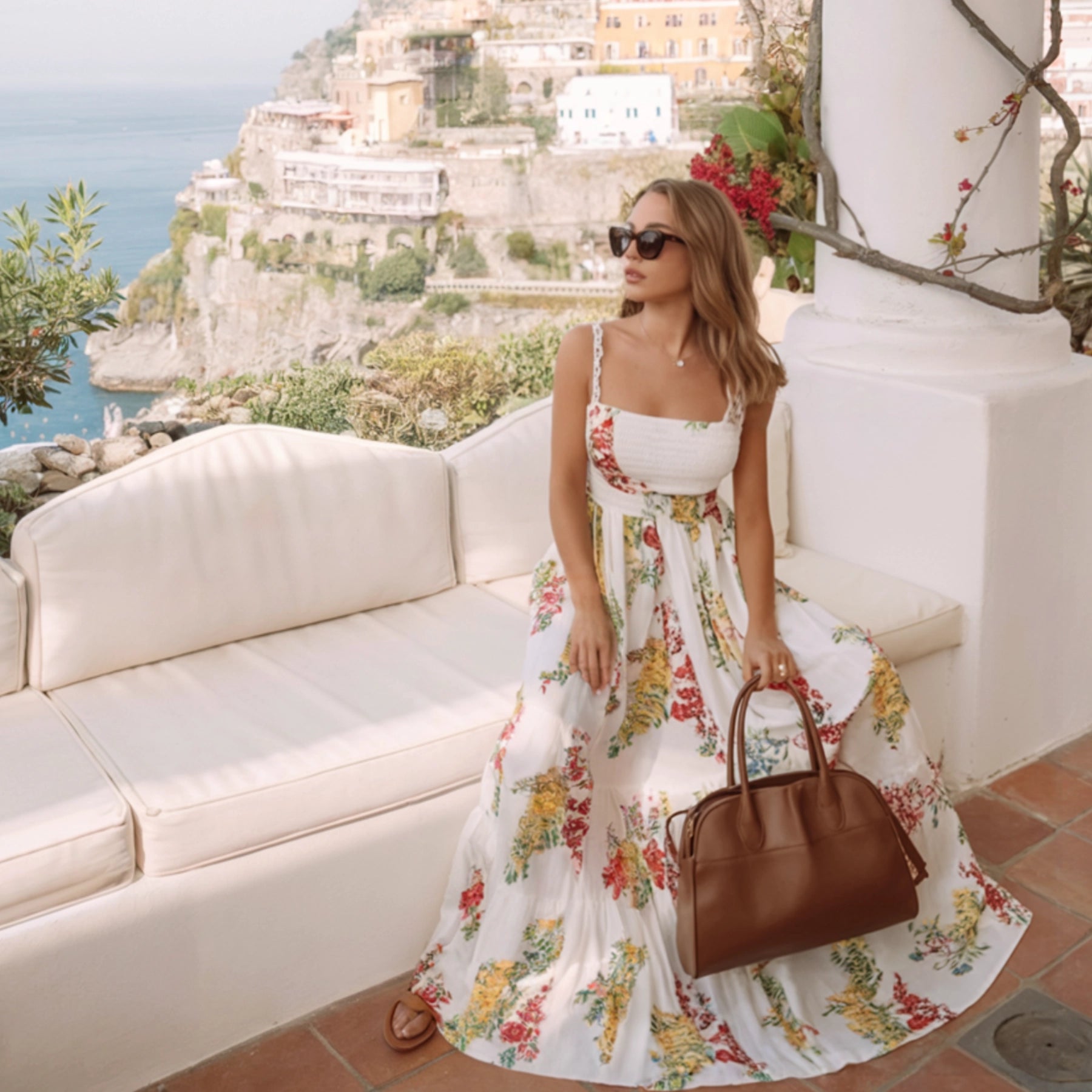 Woman in a floral dress holding a brown handbag, sitting on a white couch with a scenic background.