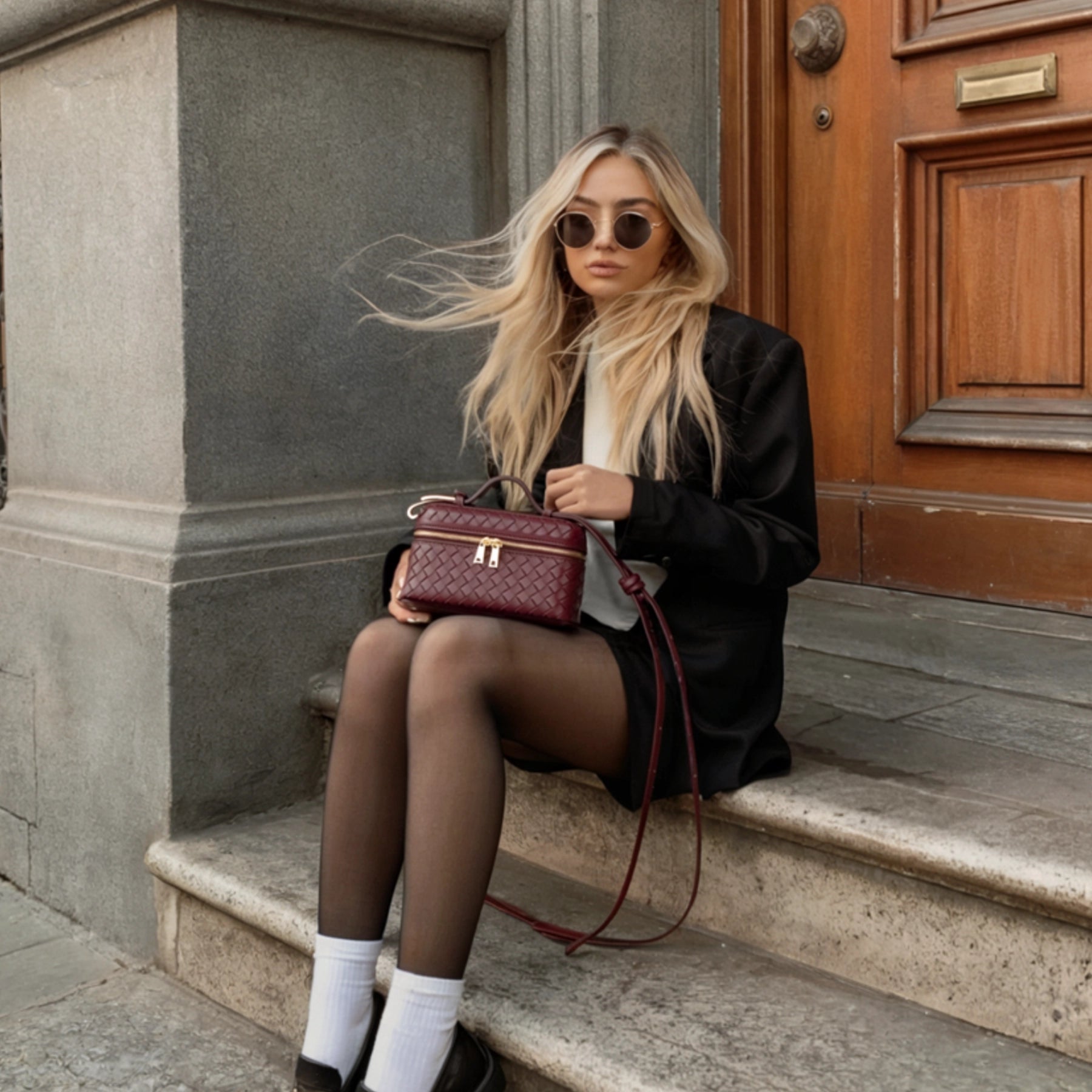 Woman sitting on steps holding a red handbag, wearing sunglasses and a black coat.