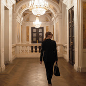 Woman in a black suit walking down an ornate hallway with chandeliers.